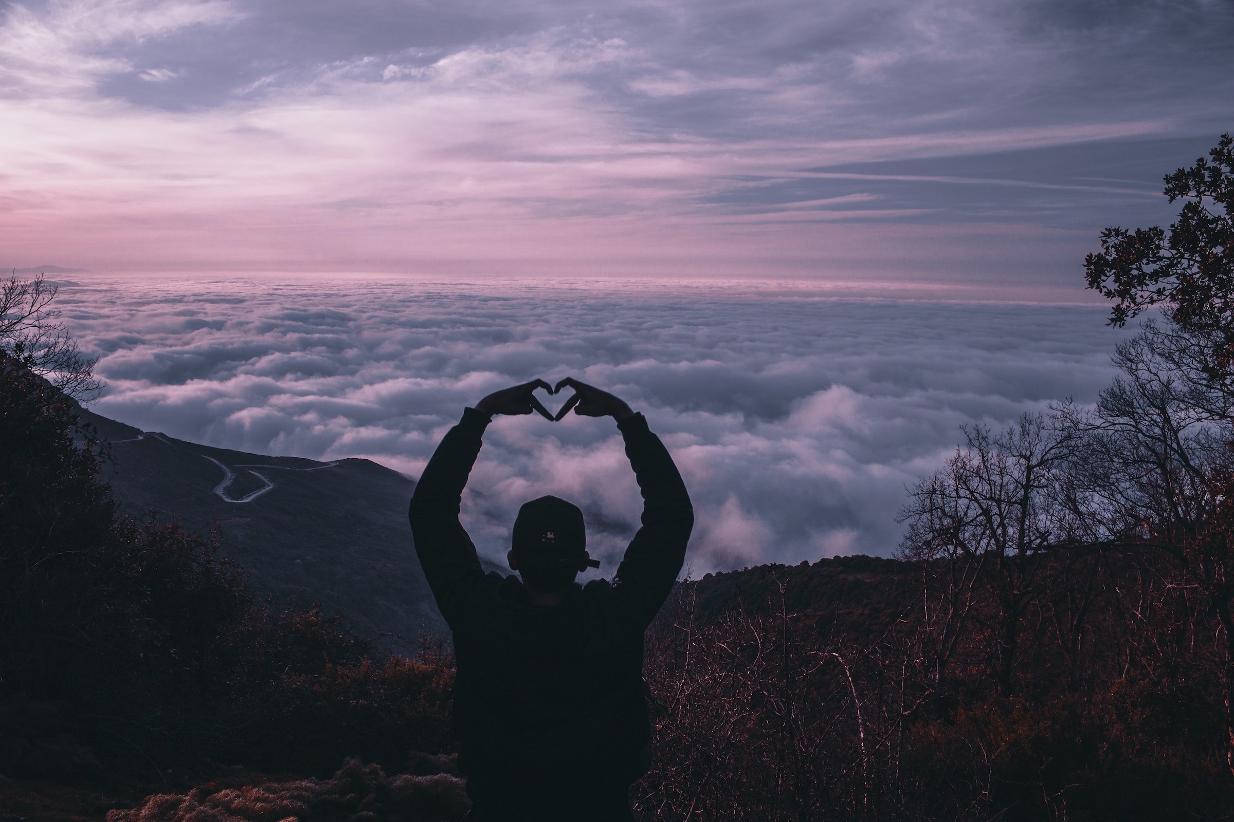 Person on a mountain creating a heart shape with hands over the clouds