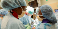 two black women gowned and masked working on a surgery in the foreground with three other people out of focus in the background.
