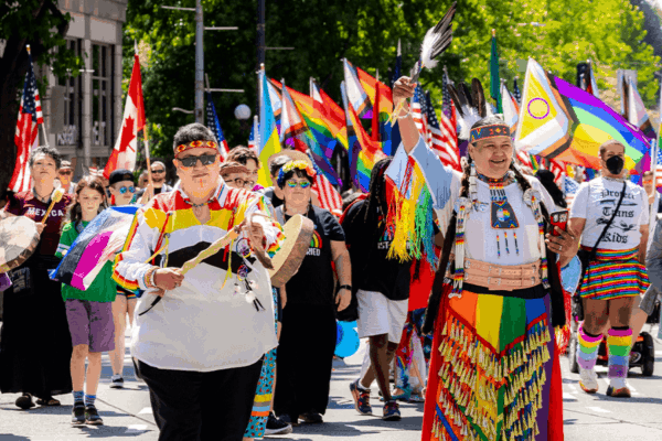 Photo of a past Pride Parade showing 2 indigenous people in the foreground with many others in the background