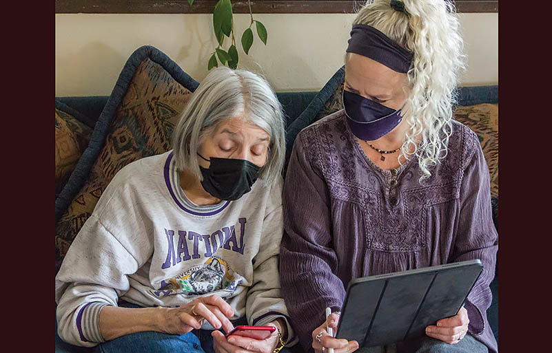 Elderly woman holds a cellphone as a younger senior woman with a tablet looks on. Both wear masks and have grey hair.