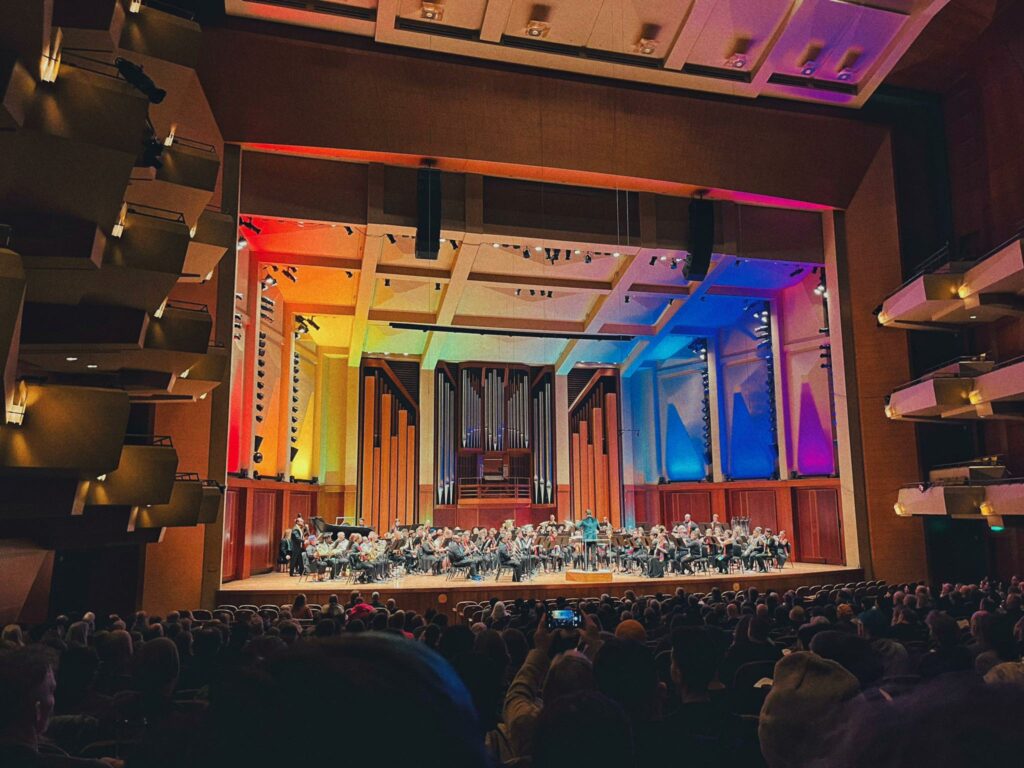 Photo of a concert stage filled with an orchestra. The stage is lit in a rainbow of color. 