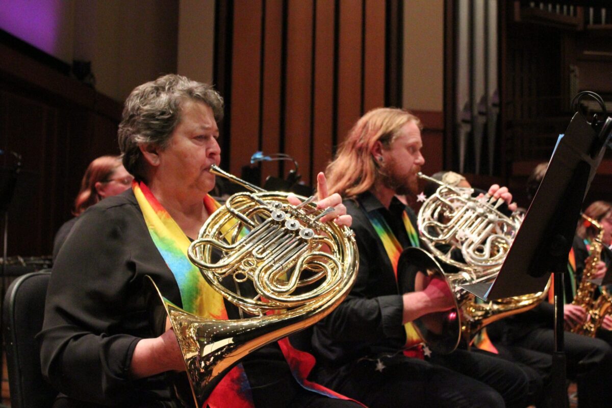 Close up photo showing two french horn players who are wearing rainbow scarves around their necks.